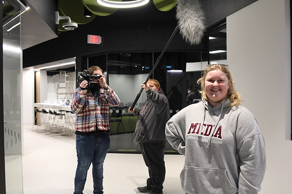 Three media students standing in the modern-looking hallway of the Centre for Creative Media, holding camera equipment.