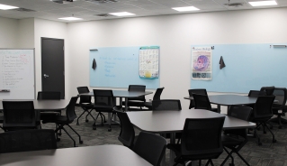 Centre for Adult Learning Brandon classroom with light-blue boards on the walls and dark grey square tables. There is a dark grey door in the far left corner, and a stand-alone whiteboard next to it.