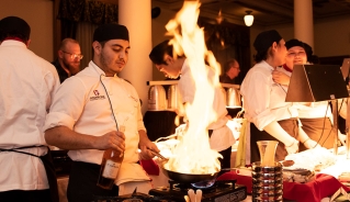 A male student in white culinary uniform holding a flaming pan with a group of other culinary students working in the background.