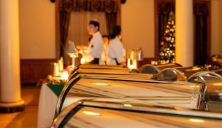 A row of closed metal food chafers lined up on the table with hospitality and culinary students shuffling around in the background and a Christmas tree in the corner of the room.