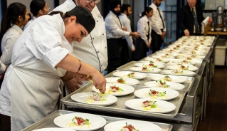 Two culinary students standing in the kitchen in front of two rows of plates with food on them, and a line of students in the background. One of them is adding finishing touches to the dish, the other one watching it, both of them smiling.