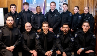 A group photo of smiling Public Safety students, with wooden paneling and Assiniboine banners behind them.
