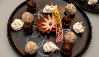 A photo of a black circular plate with a desert dish made up of various confections shaped into balls and flowers.