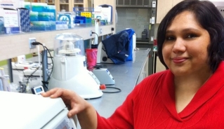 Dr. Poonam Singh standing in front of lab equipment in a red sweater, smiling at the camera.