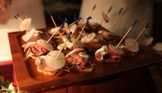 A closeup of a wooden tray of small canapes with meat, dressing, and radish slices.