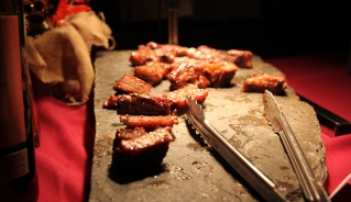 A closeup of seared pieces of meat and metal tongs placed on a flat stone, on top of red fabric, with ambient lighting highlighting the food.
