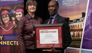 Akaps Mweeba standing in front of Assiniboine College banners with a women, holding his award certificate.