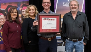 Andrew Blair standing in front of Assiniboine College banners with three other people, holding an awards certificate.