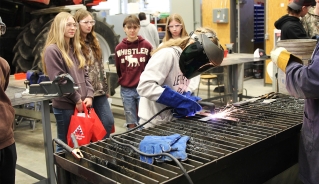 A female high school student welding a piece of metal with a group of girls standing behind her.
