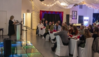 A person making a speech at the pulpit on the lit-up stage on the left side of the photo, with a crowd of guests sitting at tables and listening on the right side of the photo.