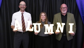 Graham Street, Madison Wright and Denis Vielfaure posing for a photo in front of a black curtain background, while holding lit up letters spelling "Alumni."