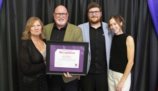 Denis Vielfaure posing with his award certificate in front of a black curtain with a group of people.