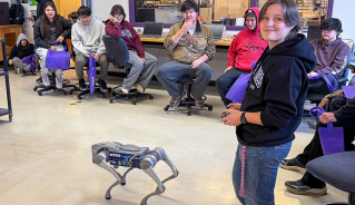 A group of high school students sitting around the room, watching a Mechatronics student operate a robot dog.
