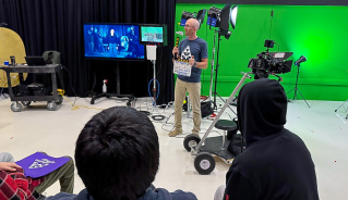 Graham street standing with a movie slate between different filming equipment, giving a presentation to high school students.