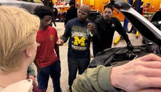 A volunteer talking to a group of students in front of an open car hood, holding an oil dipstick.