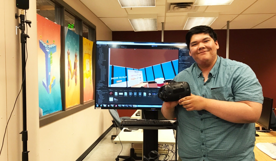 Assiniboine Alumni Dallas Flett-Wapash stands in front of a large TV monitor holding a game controler
