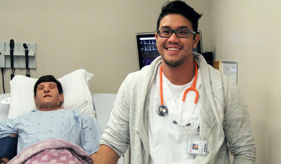 nursing student stands in front of simulation manniquin 