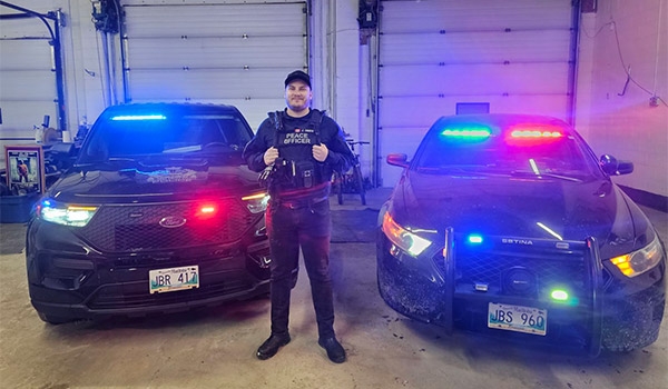 Jeremy standing in a Peace Officer uniform in front of two police cars with their lights flashing