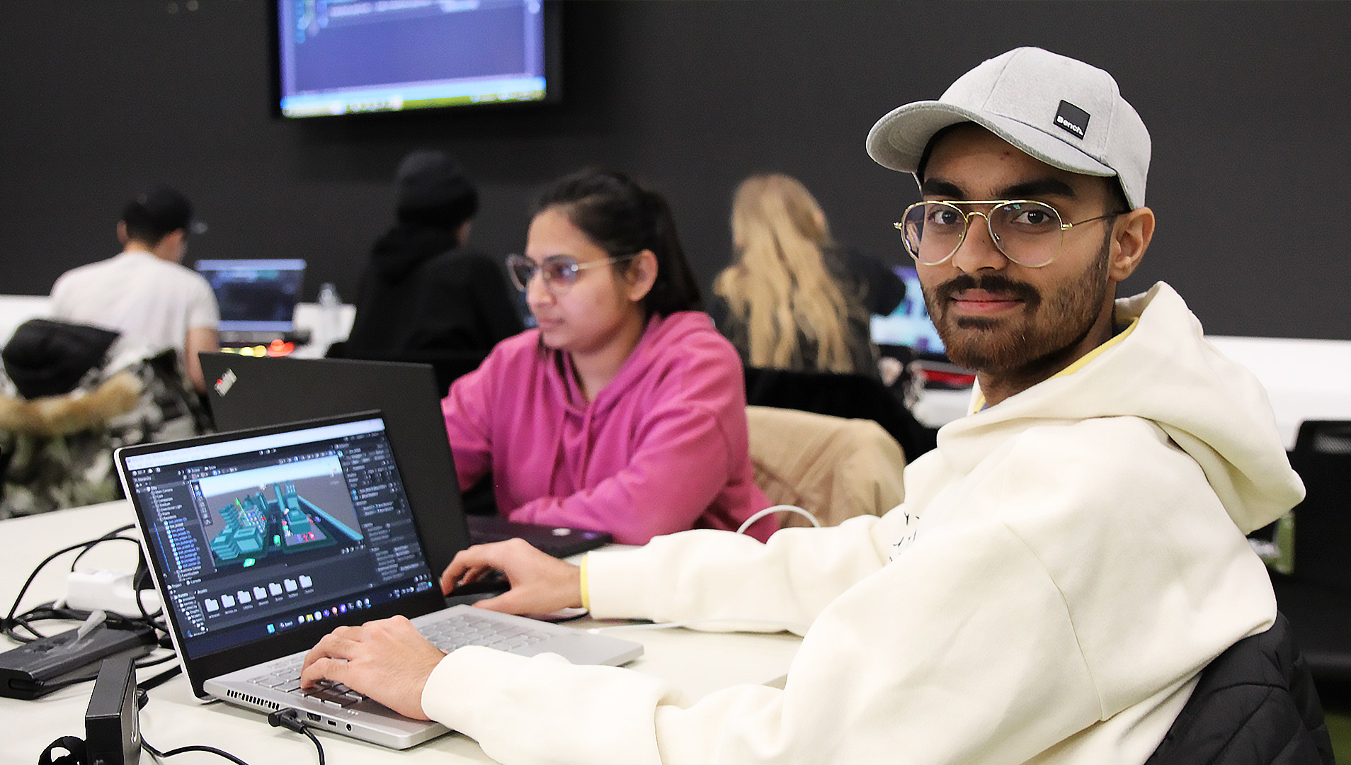 A male student sitting in front of a laptop in a classroom full of other students, working with Unity game engine.