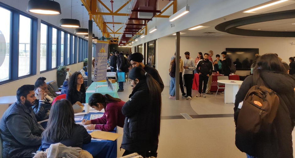 A crowd of students gathering around fair tables places along a hallway and throughout the Crossroads area adjacent to the hallway.