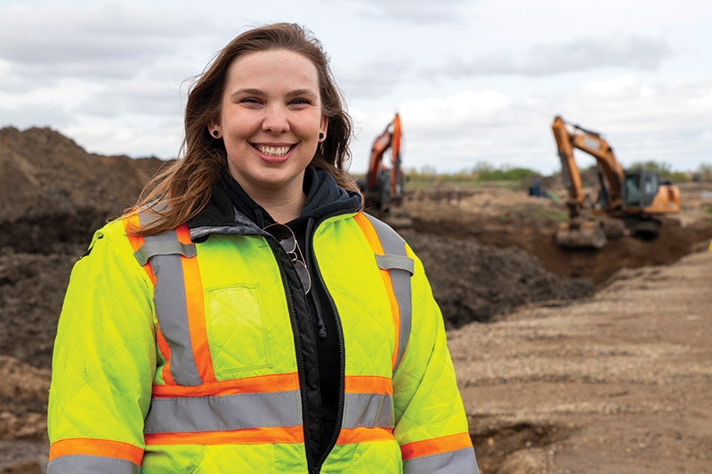 Rylee Martin wearing a high visibility vest, smiling at the camera, excavators working in the background.