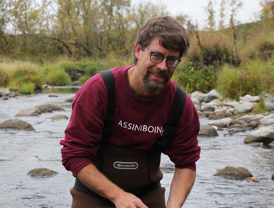 James Hood, an Assiniboine instructor, standing in the middle of a shallow river, smiling.