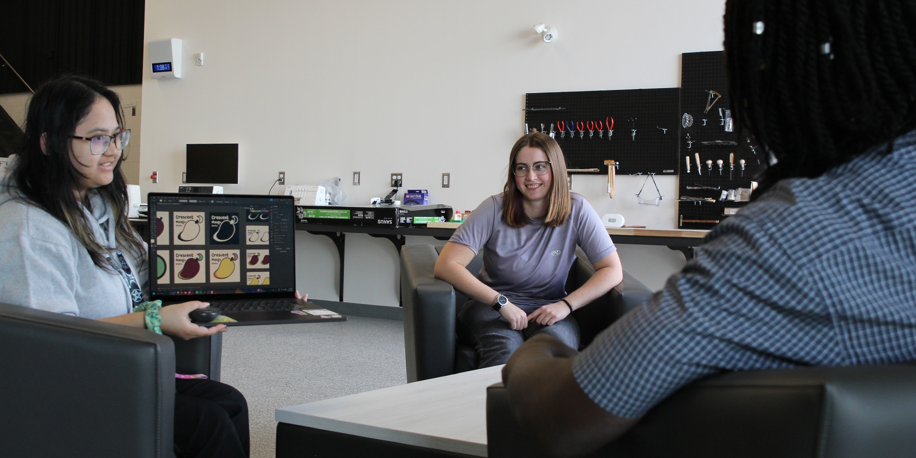 Three people sitting in armchairs around a coffee table inside the Colab, one of them showing a project on their laptop to others.