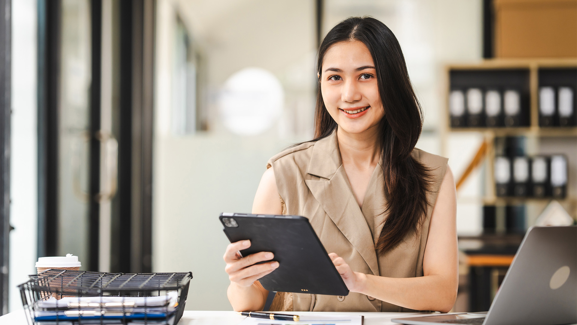 A woman with straight dark hair smiling at the camera while sitting at a desk and holding an iPad.