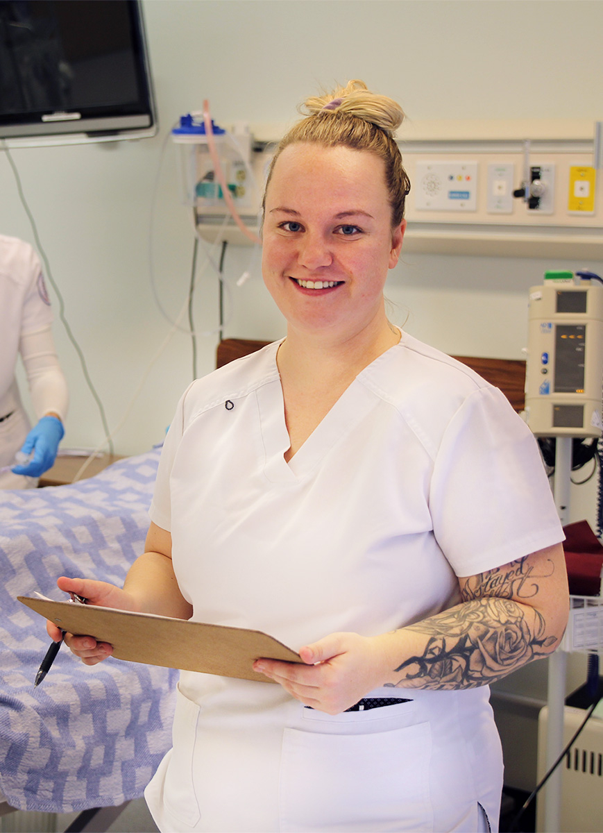 Nursing student in white scrubs standing with a clipboard in front of a hospital bed.