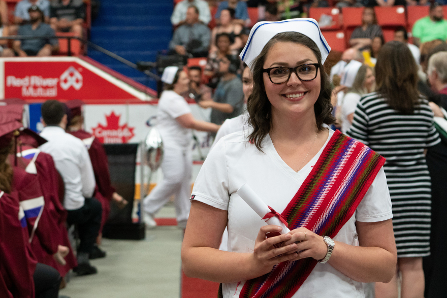 Nursing graduate stands smiling at camera holding diploma scroll.