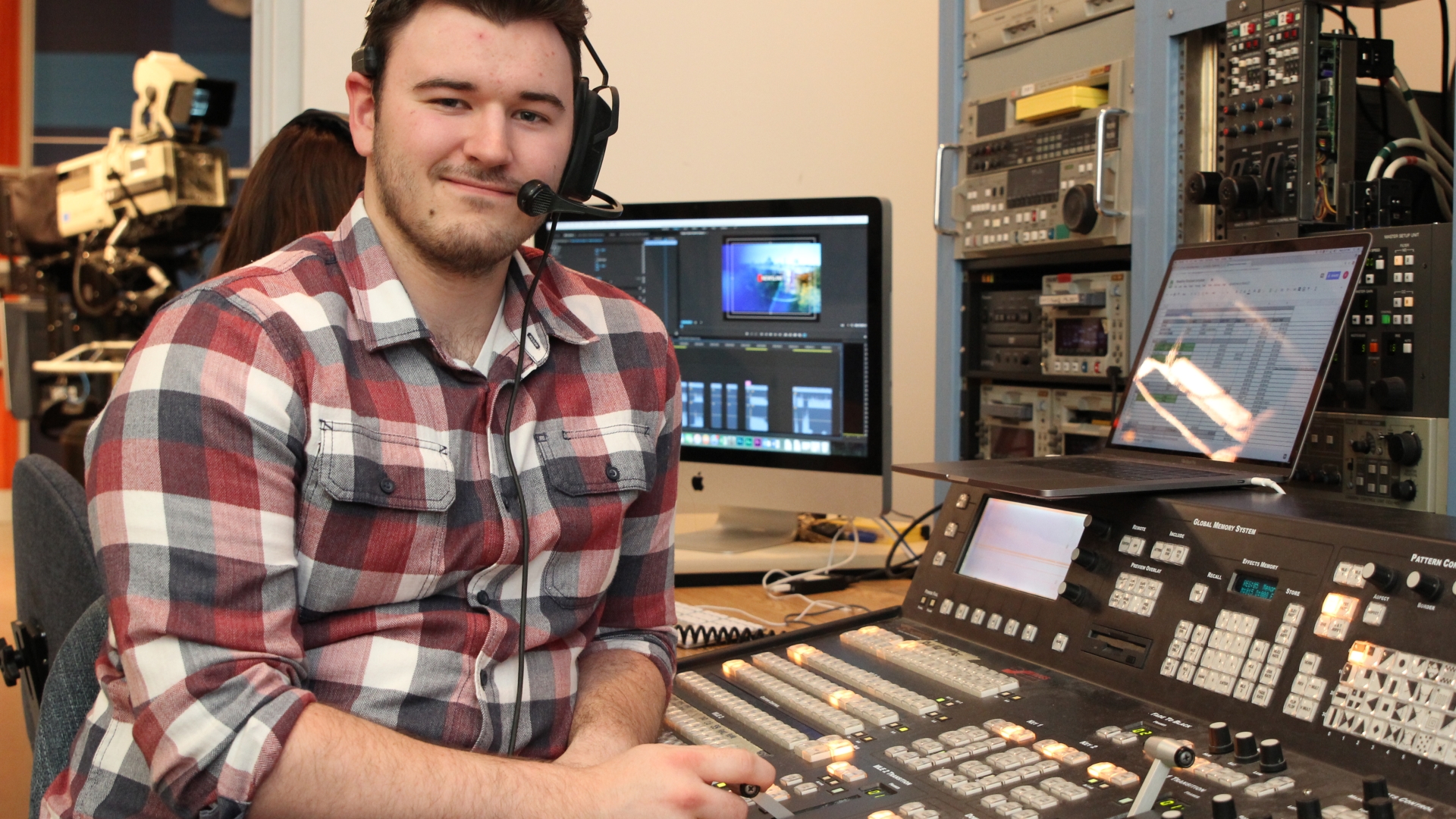 Students sits in a newsroom setting with a headset mic on.
