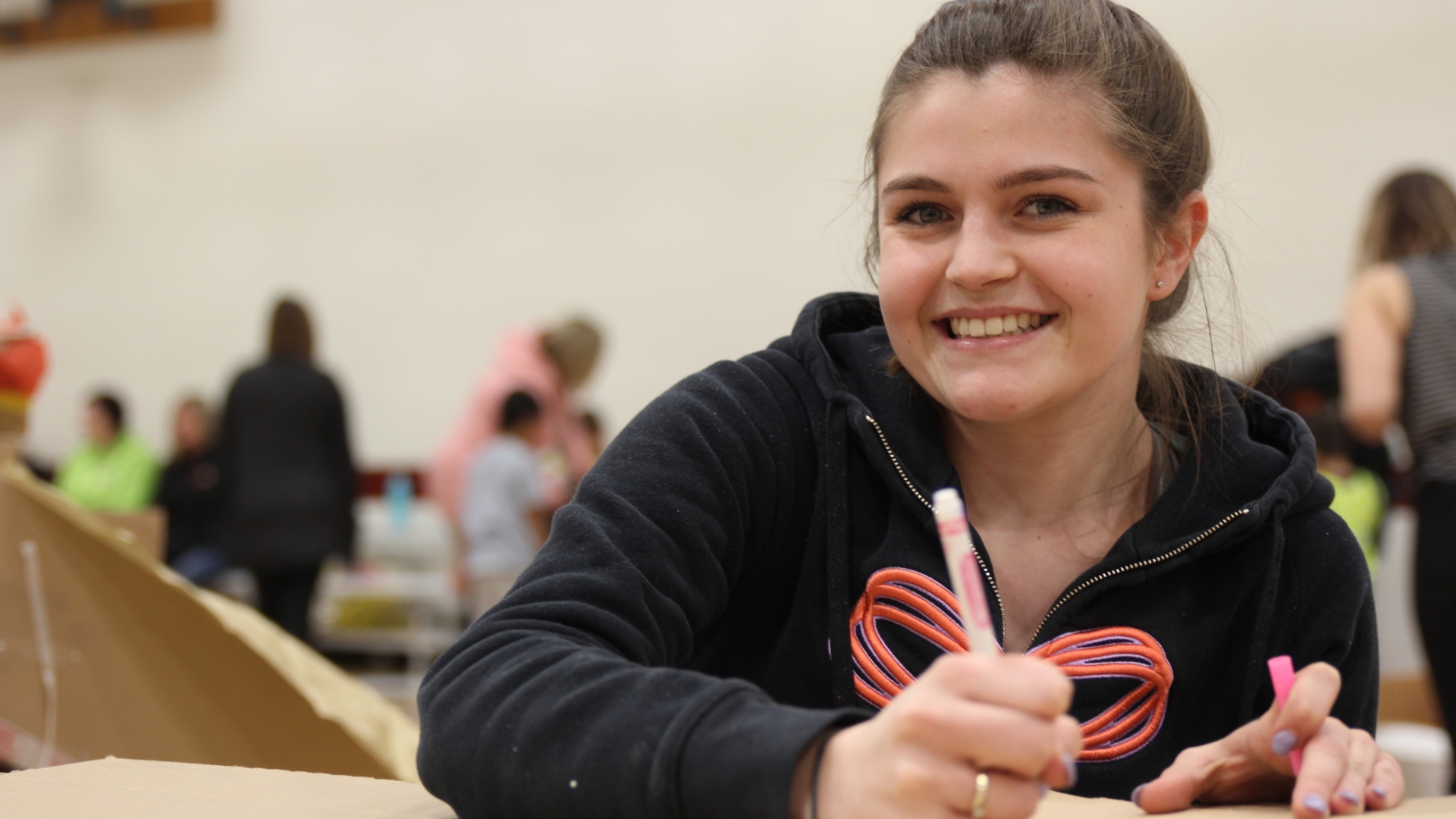 An ECE student writes with a pink marker as students play in the background