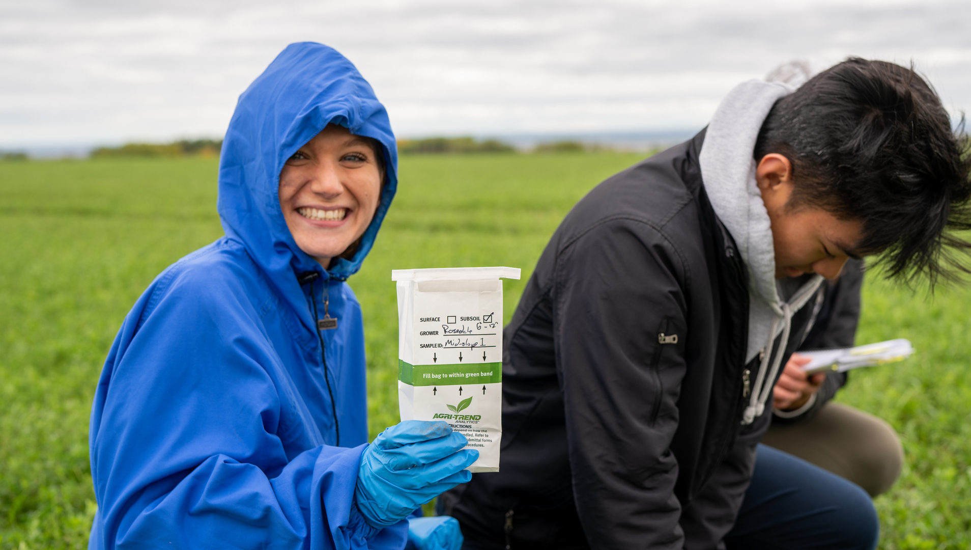 An agriculture student in a blue jacket smiles while holding up a research sample. A classmate in a black jacket looks down at the field behind her. 