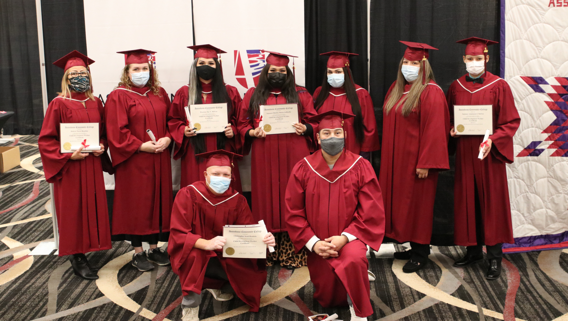 A group of nine graduates pose for a photo wearing their caps and gowns and holding their diplomas.