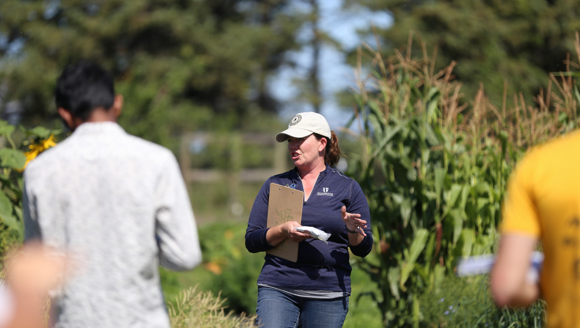 Agriculture instructor, Danielle Tichit, stands in the middle of the photo holding a clipboard. She is in focus while two students on either side of her, facing her, are blurred along with the corn crop in the background