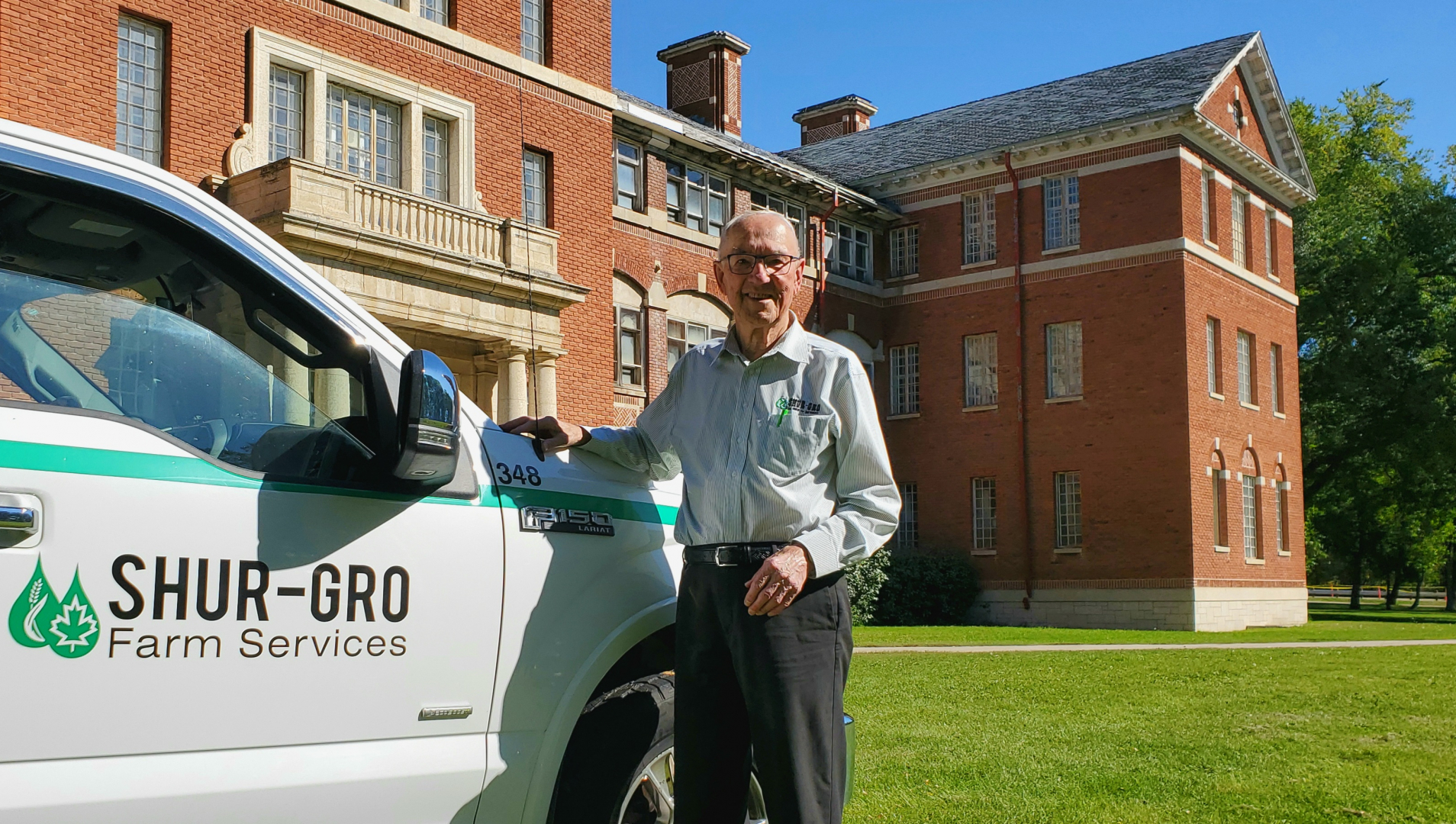 Ron Helwer stands in front of a historic building on Assiniboine's North Hill campus, leaning on the front end of his Shur-Gro Farm Services truck 