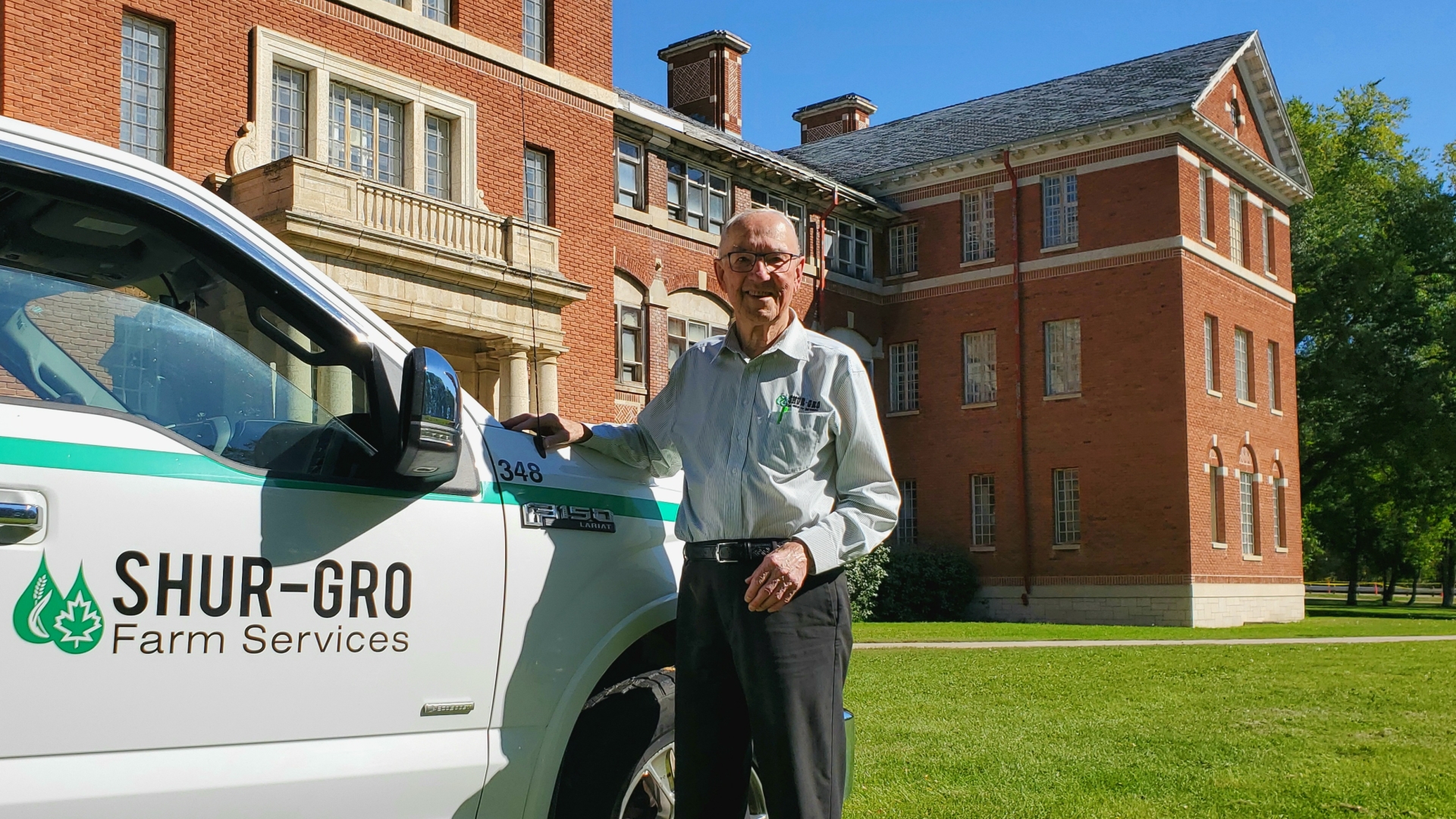 Ron Helwer stands in front of a historic building on Assiniboine's North Hill campus, leaning on the front end of his Shur-Gro Farm Services truck 
