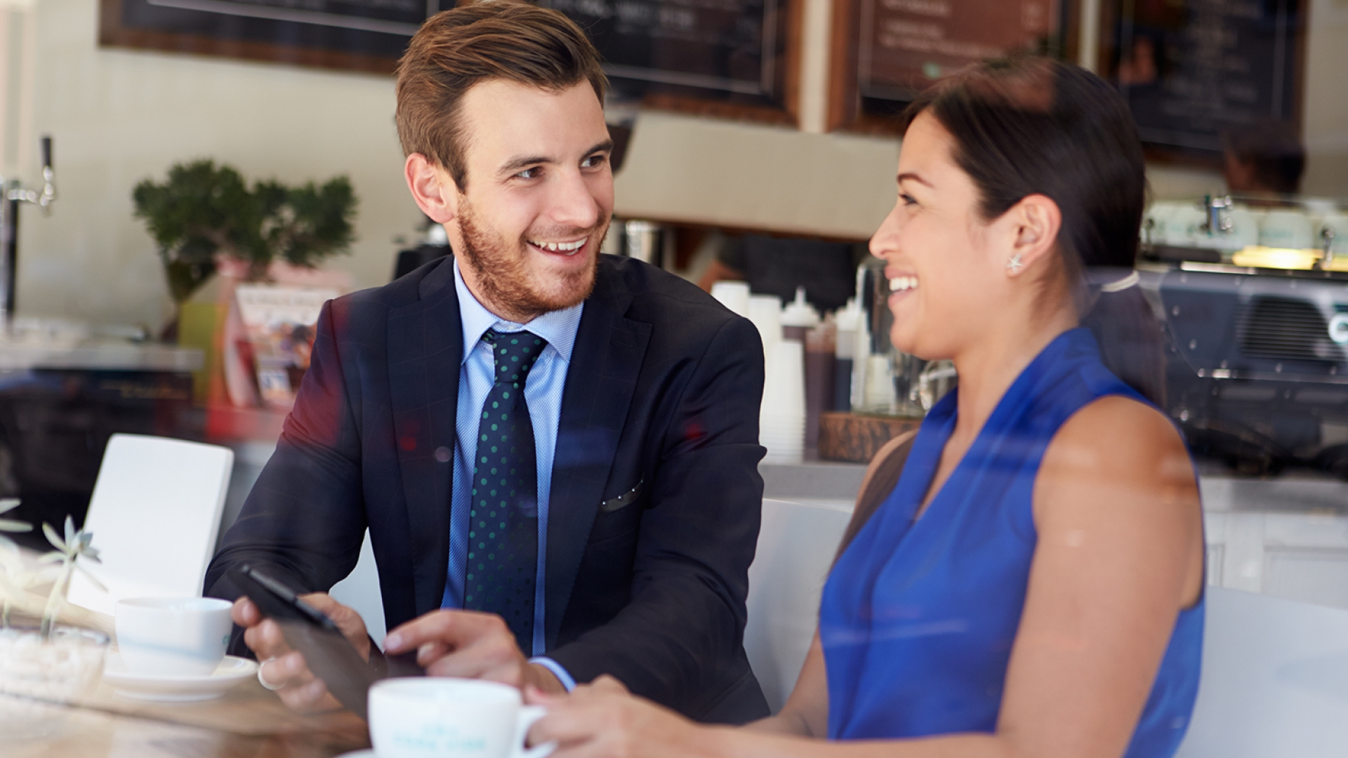 Photo is shot through a window into a coffee shop. A man with short brown hair faces the window in the left of the photo, looking at a woman in a blue tank top with a long dark ponytail the right of the photo, showing her something on a tablet. 