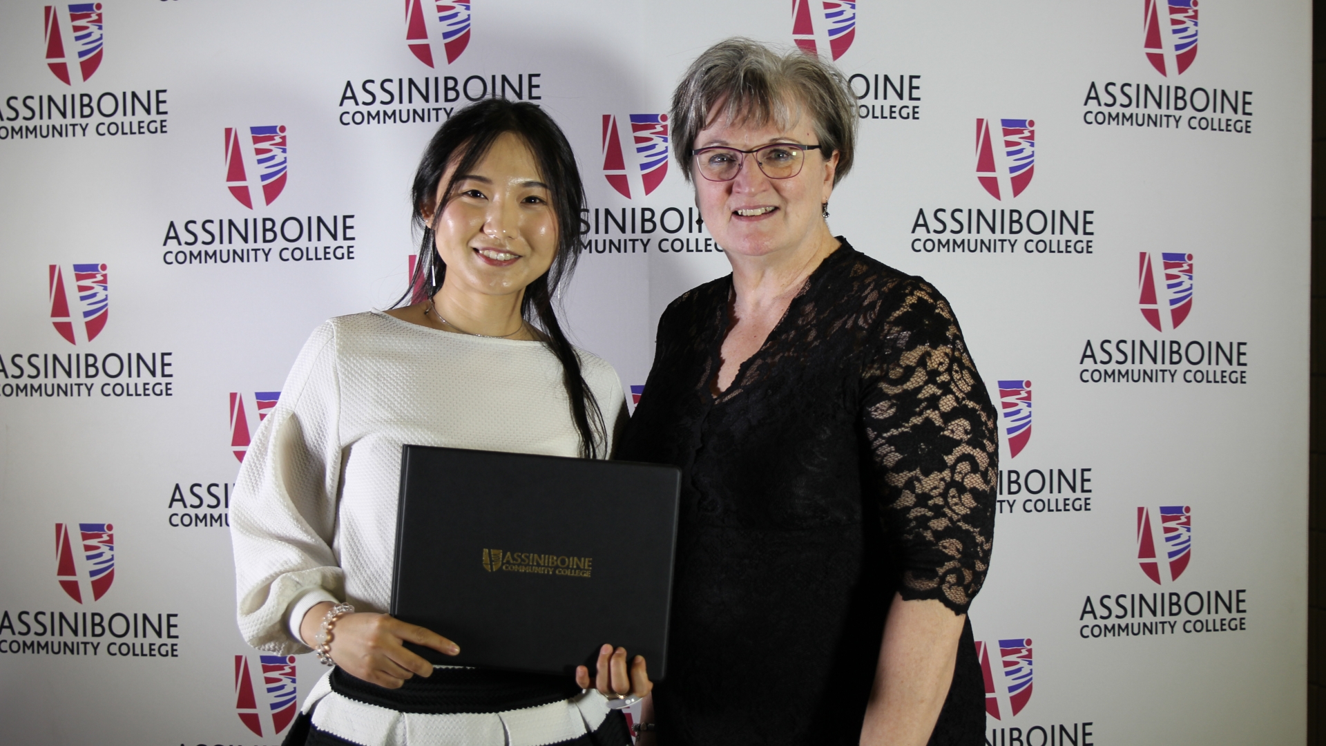 Two women pose facing the camera - a student and the Dean of Business. The student, in the left of the frame holds a certificate encased in an Assiniboine Community College presentation folder. 