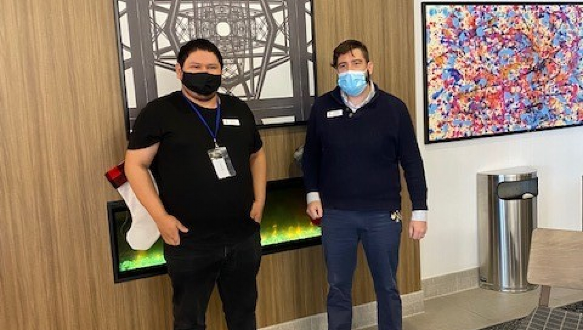 Two men stand in masks, in front of a wall with a piece of art behind them.