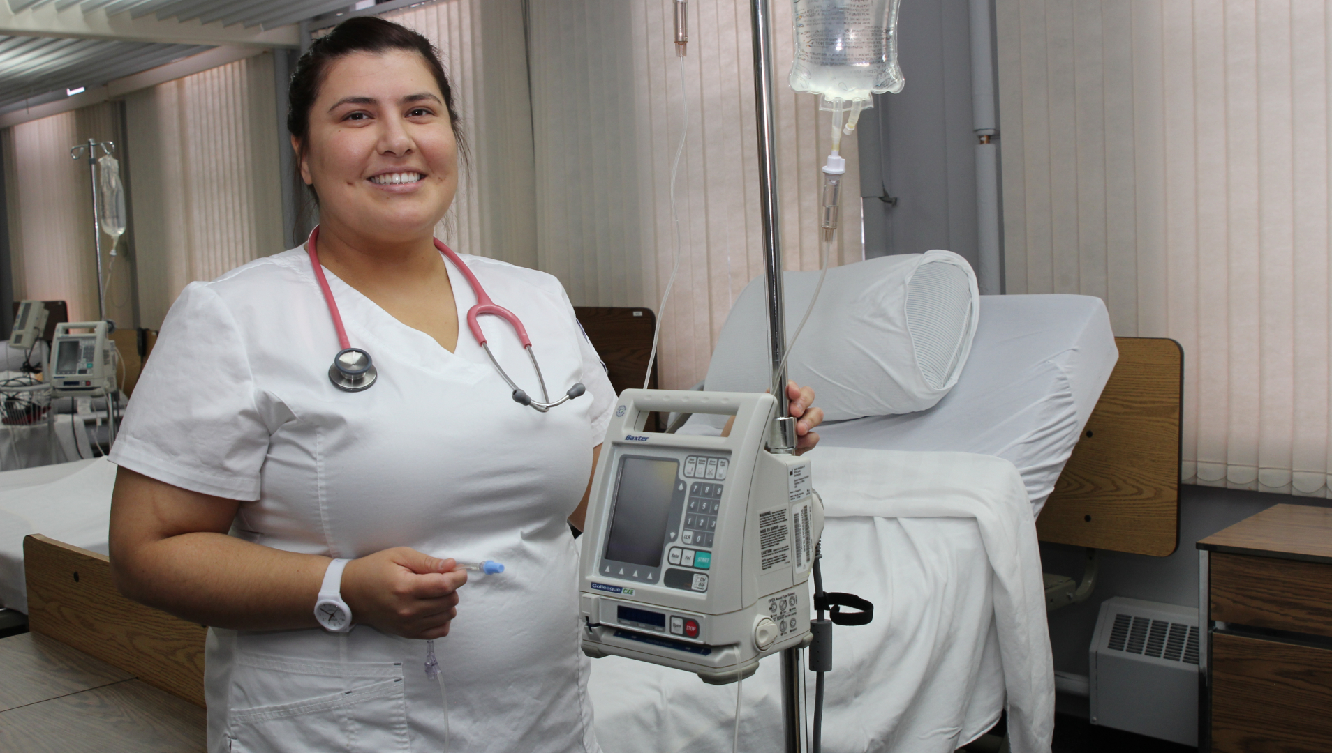 A nursing student stands with an IV pump in hand in front of an empty bed 