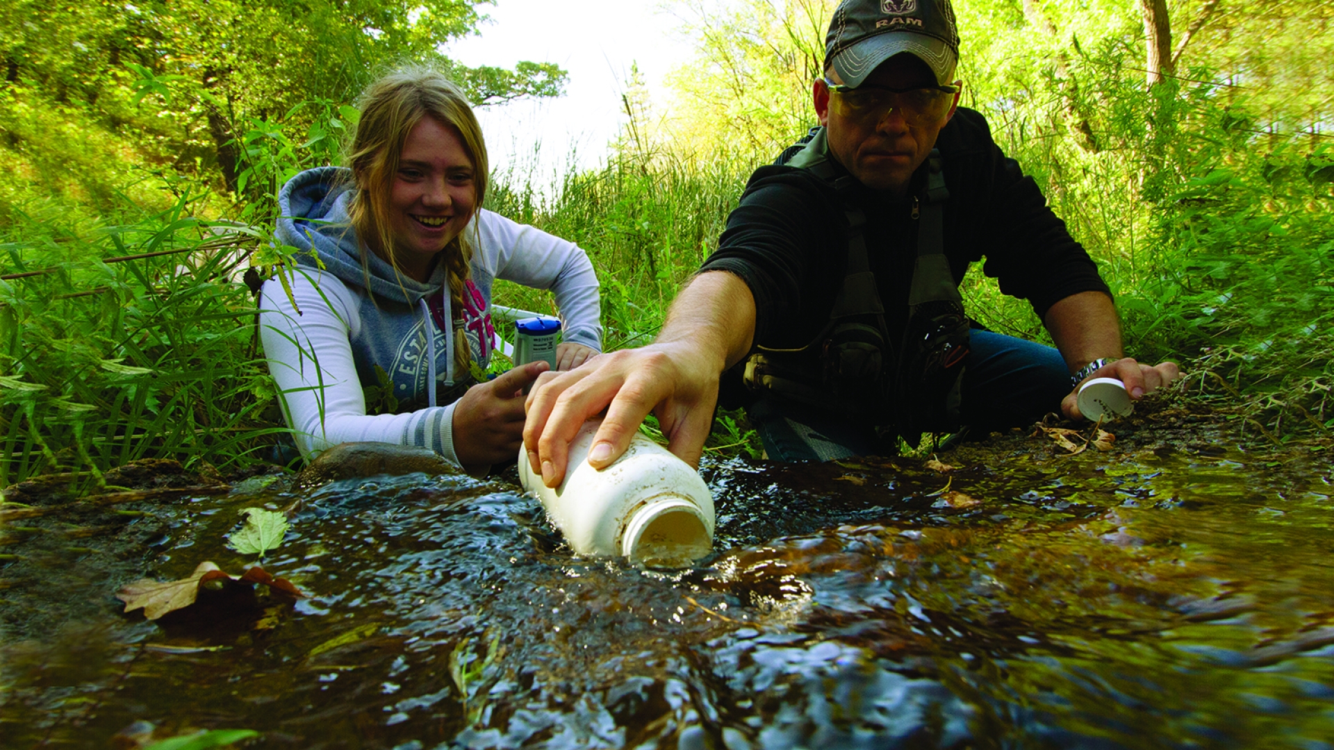 A student on the left, kneels above a creek beside an instructor on the right collecting a water sample