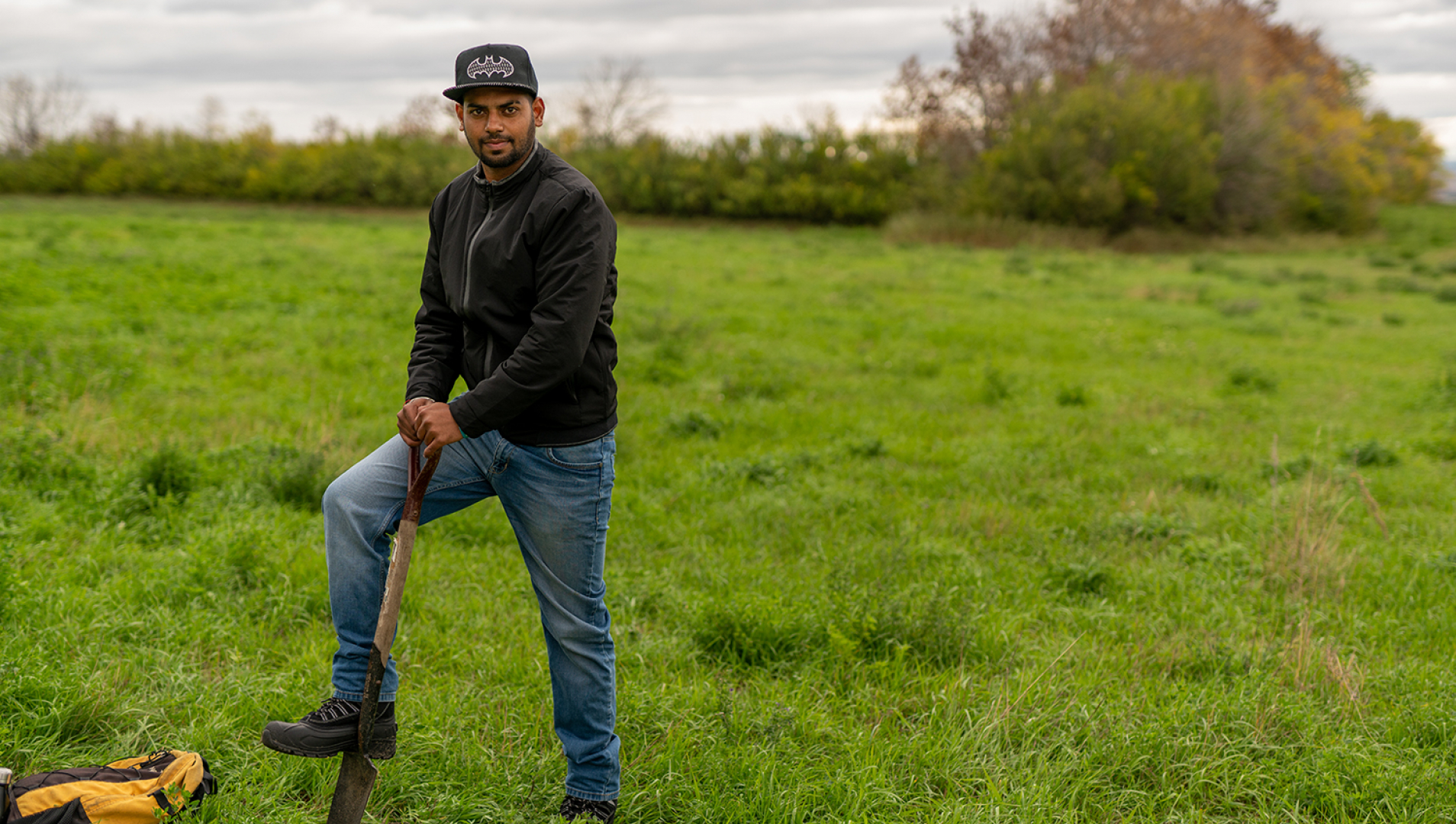A student stands in a field in the right third of the frame with his foot on a spade digging into the ground.