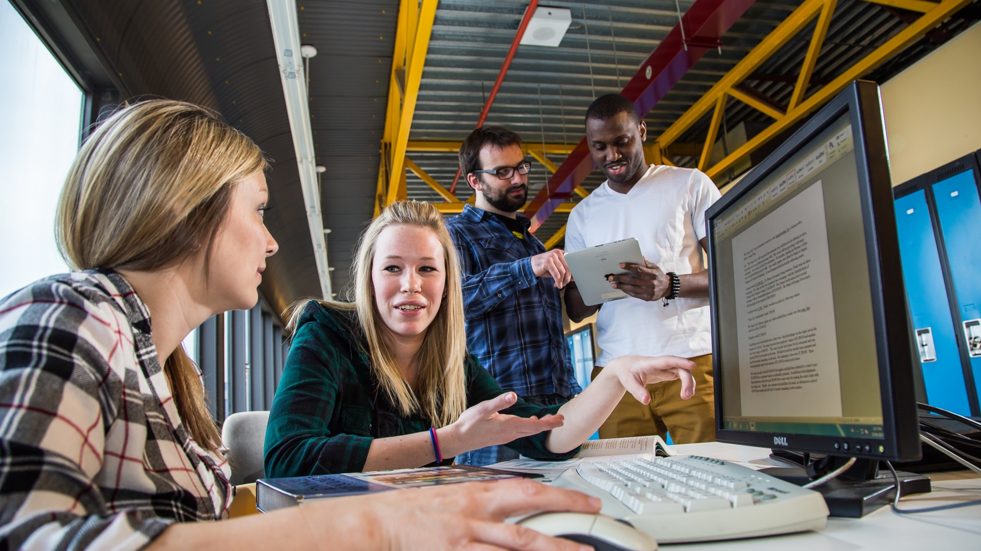 Two female students in the bottom left of the frame sit at a computer and appear to be discussing something. In the mid-right top of the frame behind them, two male students look at a device together. 