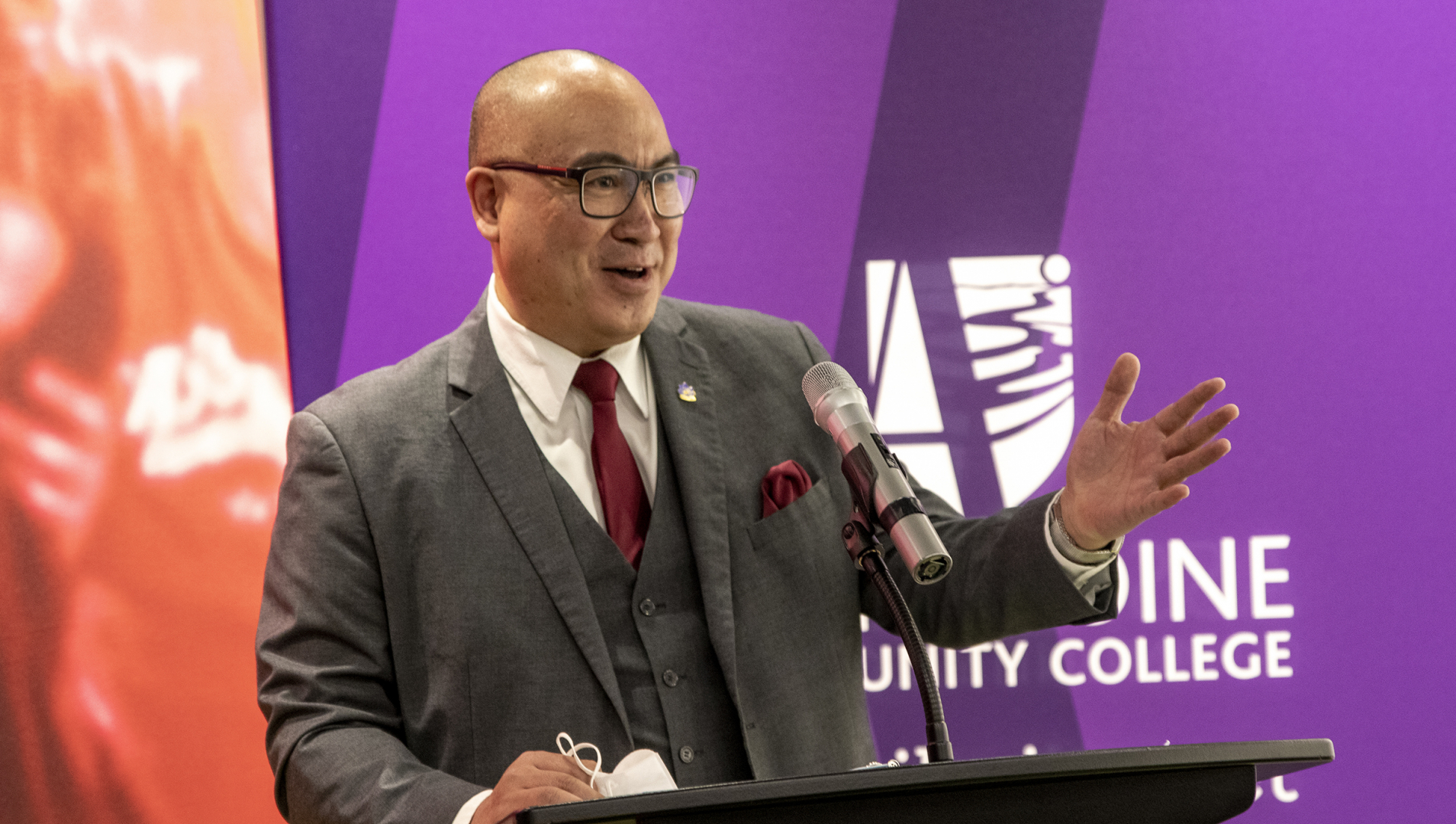The Honourable Jon Reyes, Minister of Advanced Education, Skills and Immigration stand at a podium, speaking into a microphone and gesturing toward the crowd out of frame. 