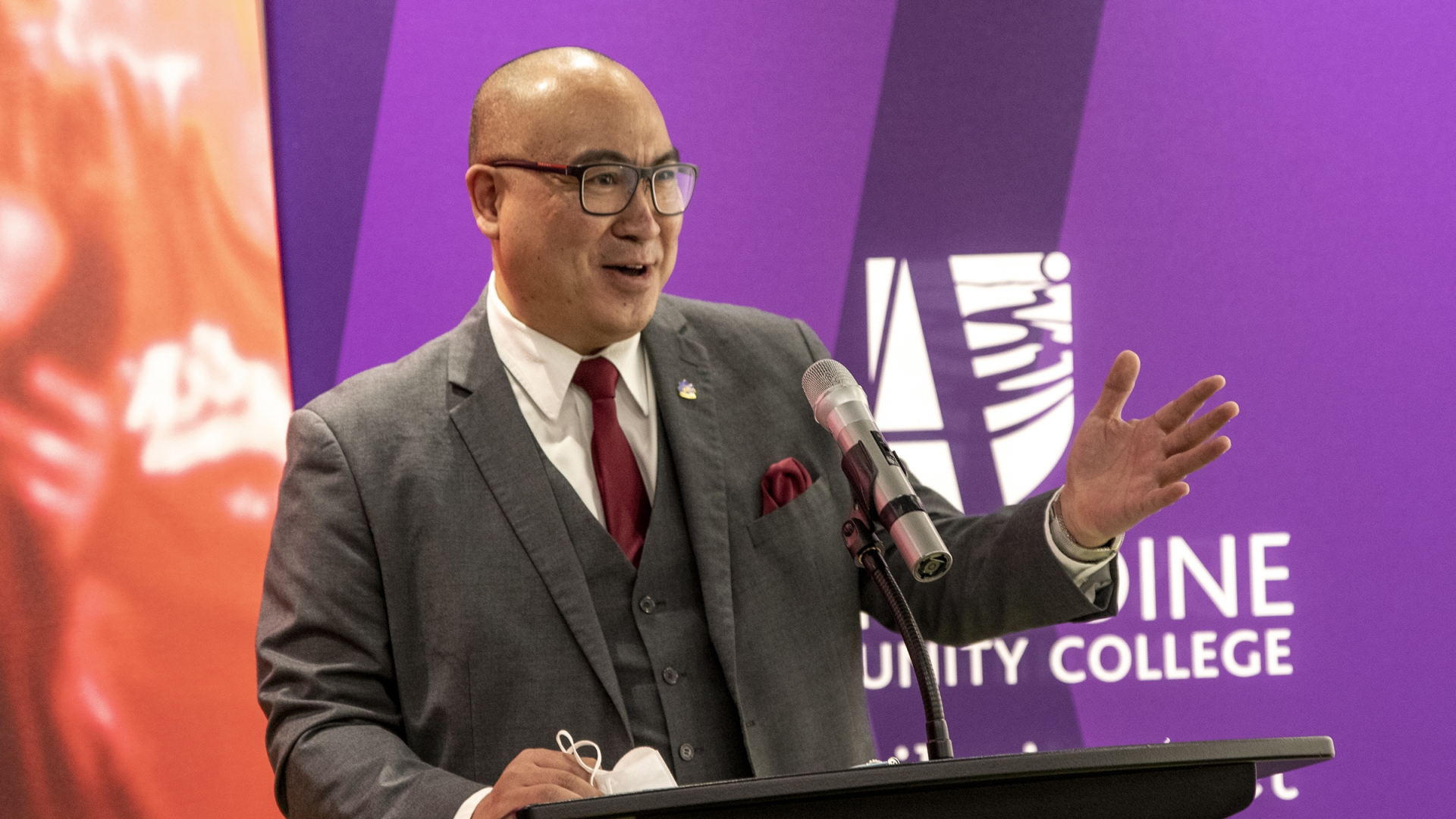 The Honourable Jon Reyes, Minister of Advanced Education, Skills and Immigration stand at a podium, speaking into a microphone and gesturing toward the crowd out of frame. 