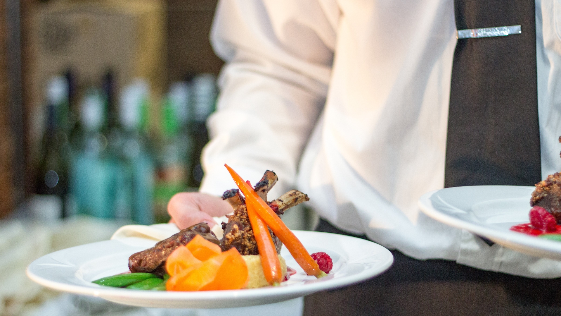 A plate of food in the bottom left of the frame is held by a student whose torso is visible in the background. The student wears a black tie on a white shirt and the food appears to be carrots and other vegetables with beef in the middle of the plate. 