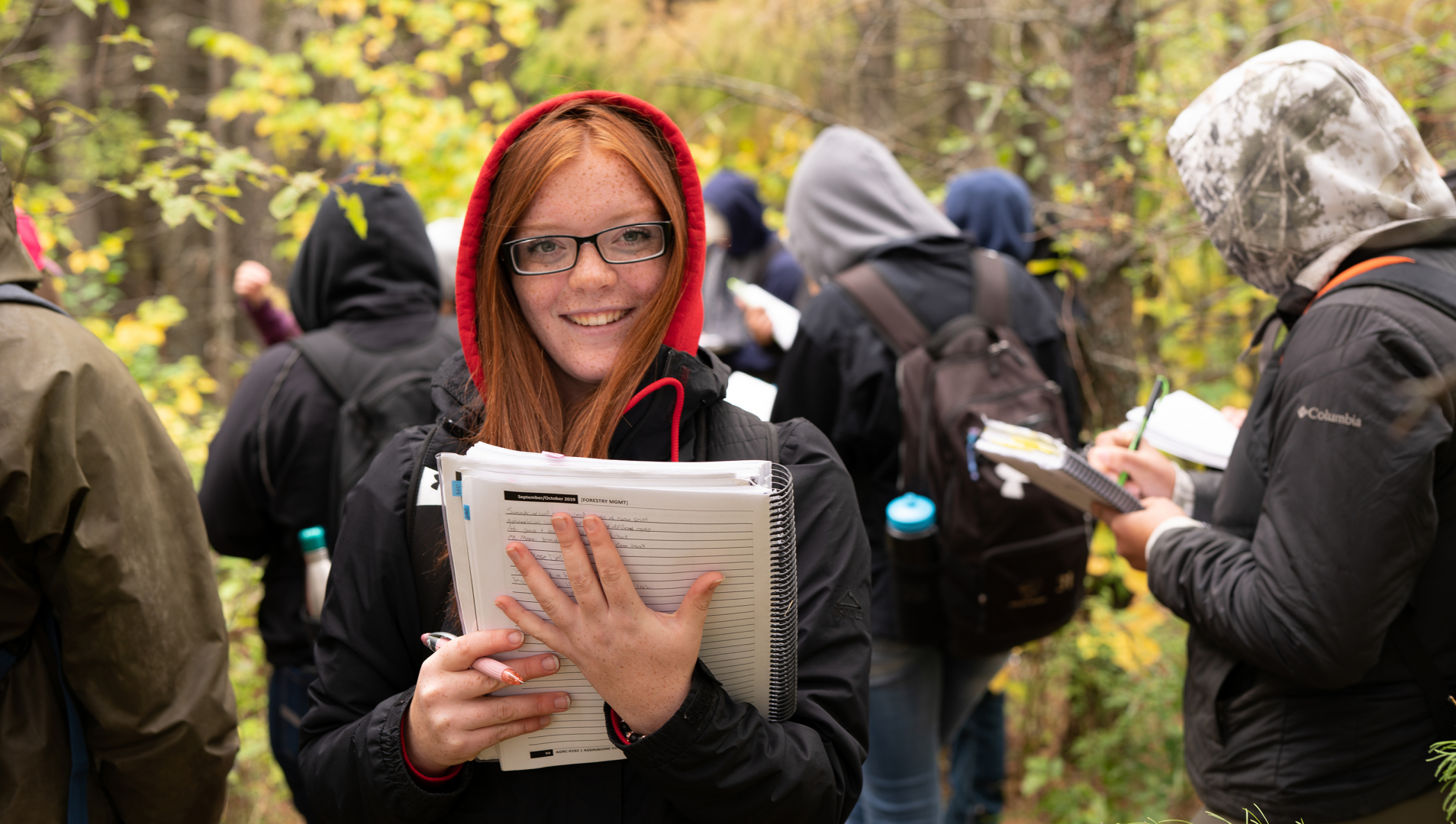 A student with long red hair and a red hood up on her head looks at the camera and holds a notebook. The student stands amongst trees and other students in the background.