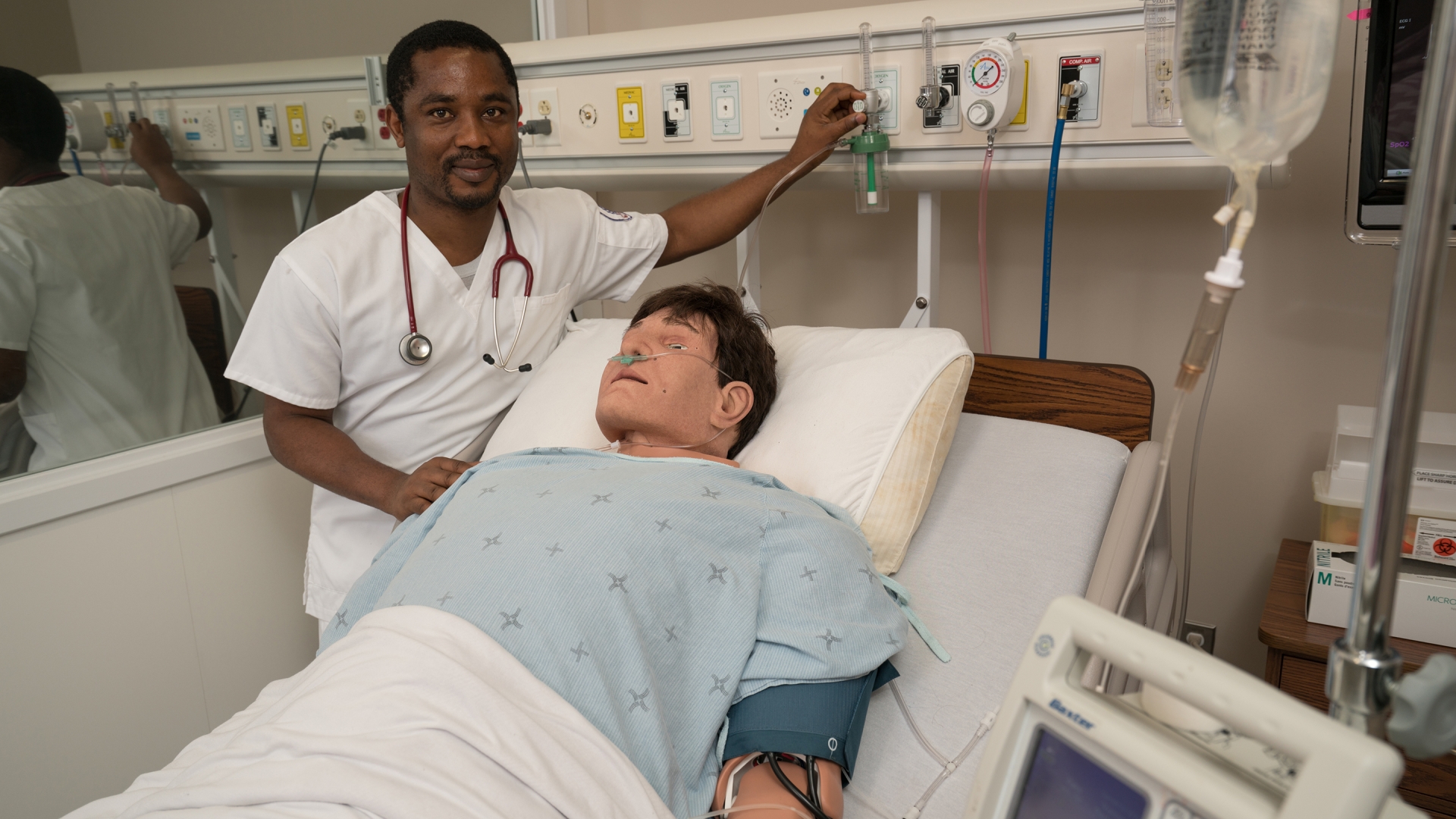 A nurse stands in the left of the frame to the left of a hospital bed with a simulation patient lying in the bed.