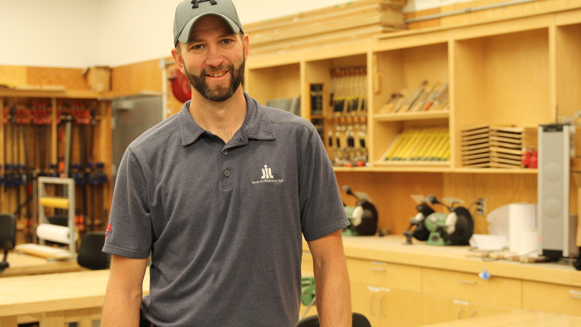 Josh Church stands in the left of the frame, wearing a grey polo t-shirt, dark pants and a grey cap. In the background is his carpentry shop.